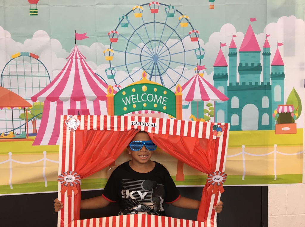 Boy wearing shades appears in theater prop as if on stage. Backdrop is ferris wheel and big top.